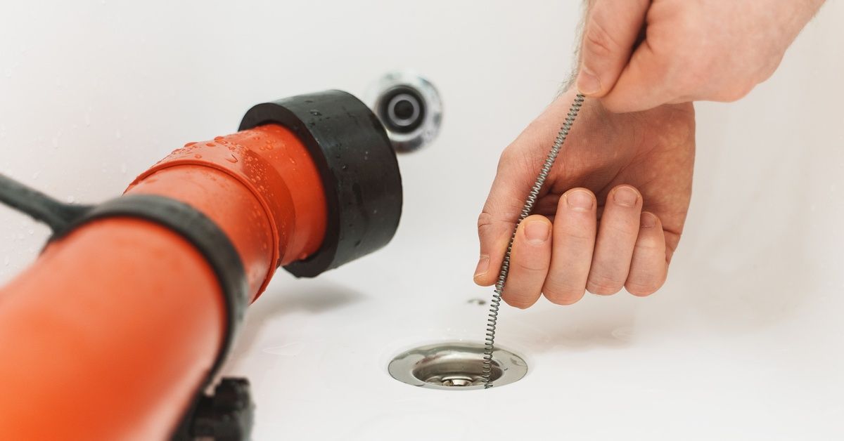 A pair of hands guiding a snake drain into the drain of a bathtub. There is a bright orange tube sitting next to the drain.