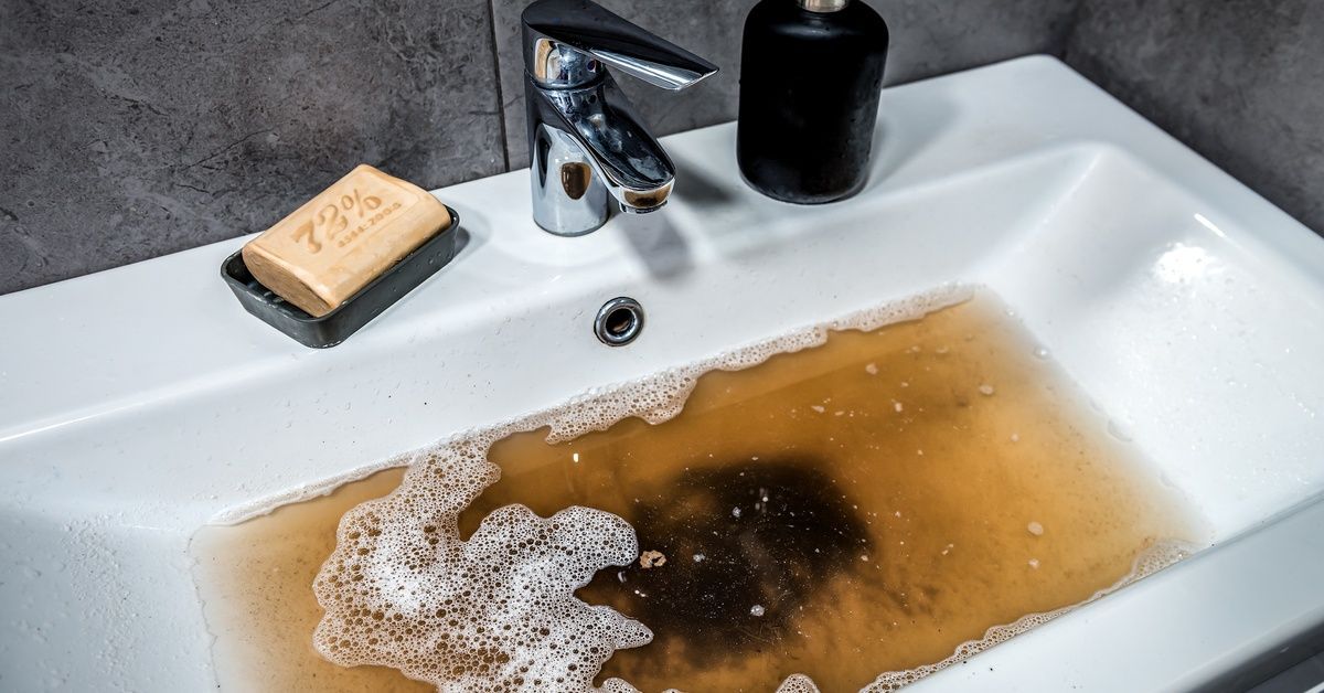 A white bathroom sink filled with dirty brown water and soap residue pooled around the drain from a clog.