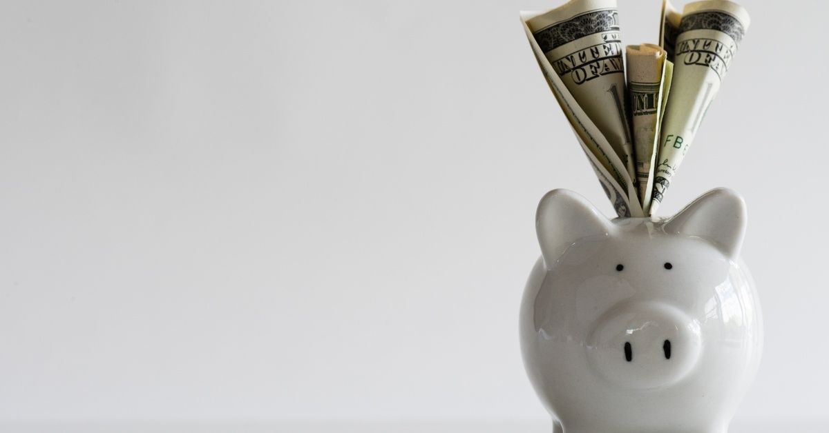 A white piggy bank with US dollar banknotes stuffed inside and sticking out of the slot on a white surface.