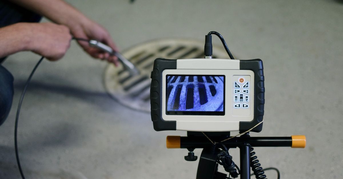 A man feeding a sewer inspection camera cable into an indoor floor drain while kneeling.