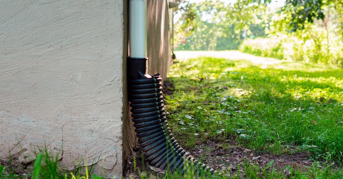 The side of a house with a drainage downspout running down it. At the end of it is a flexible tube l