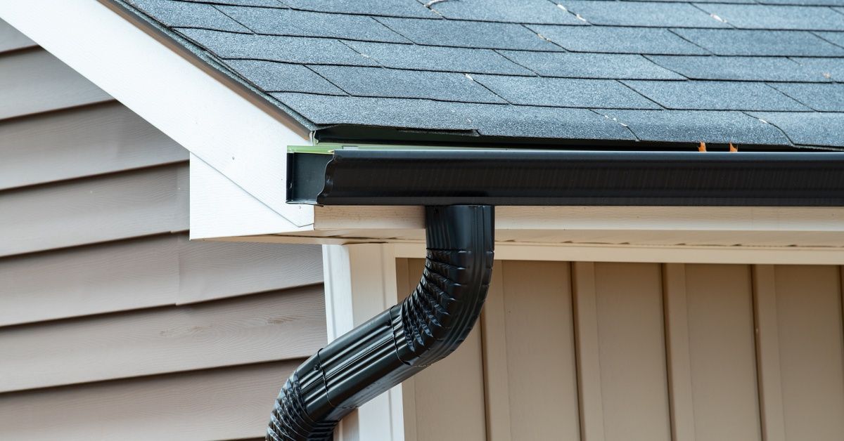 The corner of a house's roof that has a gutter connected to it. There is a downspout coming down from it.