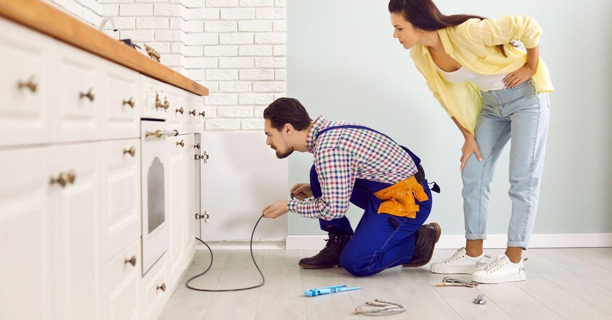 A plumber crouched next to a sink using a long, thin tube. A woman is standing behind him, watching 