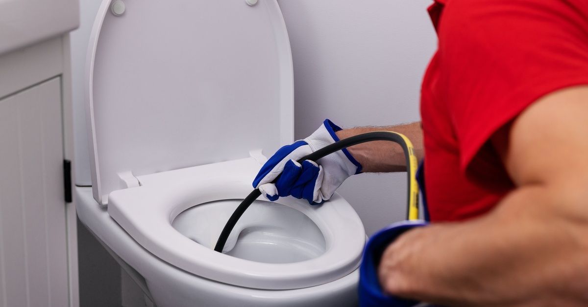 A man moving a long hose down a toilet to unclog the pipes. He is wearing blue and white gloves.
