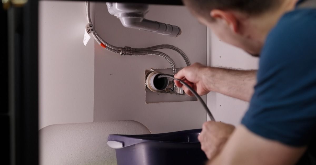 A plumber uses a spring tube tool to drill through clogged sink pipes beneath a bathroom sink basin