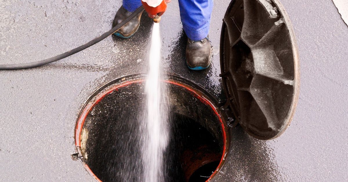 A person wearing blue pants stands over an open drain cover, spraying pressurized water into a hole.