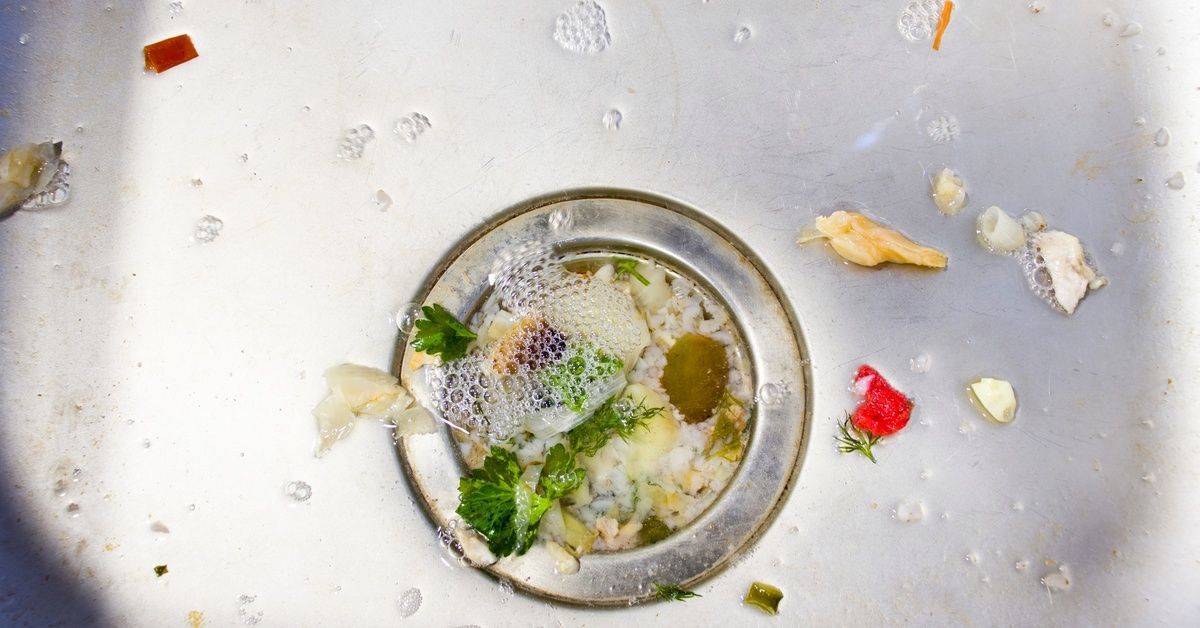 A close-up of a kitchen sink with various foot particles stuck in the drain to the point where the water is pooling.