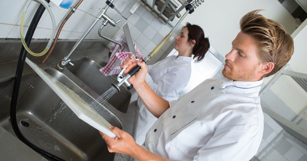 A kitchen staff works at a commercial sink station, washing cutting boards and drying dishes.