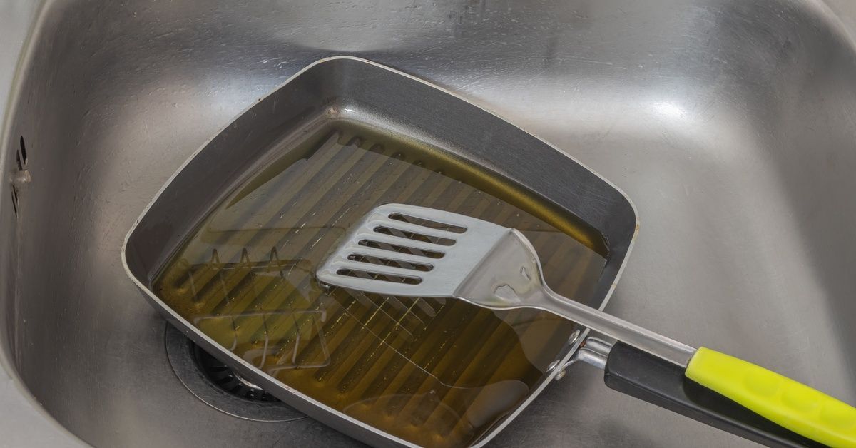 A frying pan filled with a layer of grease and a spatula rests in a gray stainless steel kitchen sink.