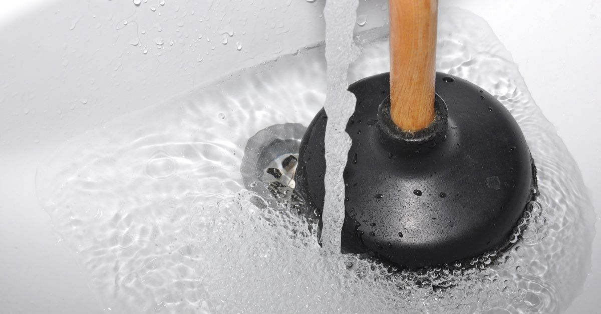 A plunger with a black rubber head resting in a white sink with the water running over a drain.