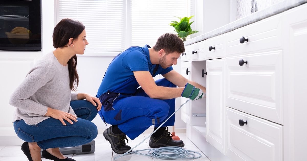 A plumber in a blue uniform and a woman kneel in front of an open sink cabinet. The plumber feeds a tool into the space.