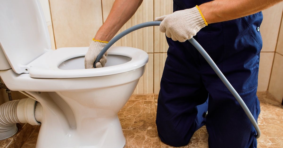 A close-up of a man kneeling down in front of a toilet, feeding a long gray hose into the toilet bowl and drain.