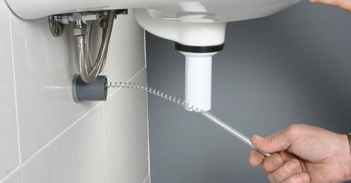 A close-up of a person's hand using a metal drain snake. They're inserting it into a gray pipe.