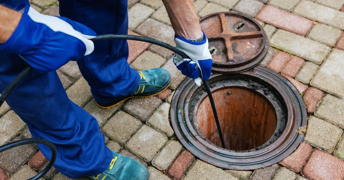 A person wearing blue pants and gloves uses a hydro jetting tool on a drain in the ground. The tool is deep in a hole.