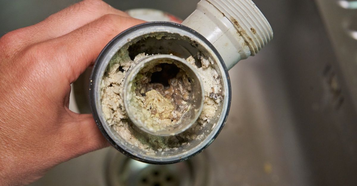 A hand holds a clogged sink pipe above a stainless steel sink.