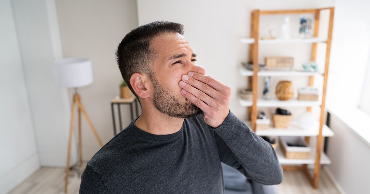 A man in a long-sleeved dark gray shirt scrunches his face and holds his nose while standing in an apartment.