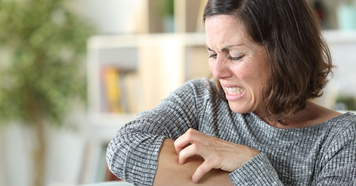 A woman in a gray ribbed long-sleeved shirt grimaces as she scratches at the skin on her right arm.