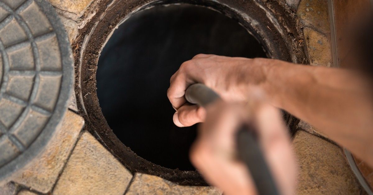 A pair of hands snakes a tube into an outdoor drain. There are brown tiles surrounding the drain.