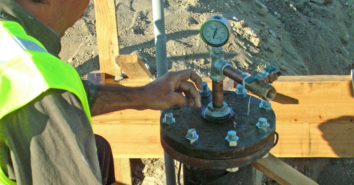 A ground-level shot of a plumber conducting a camera inspection on a pipe. The drain is located outside.