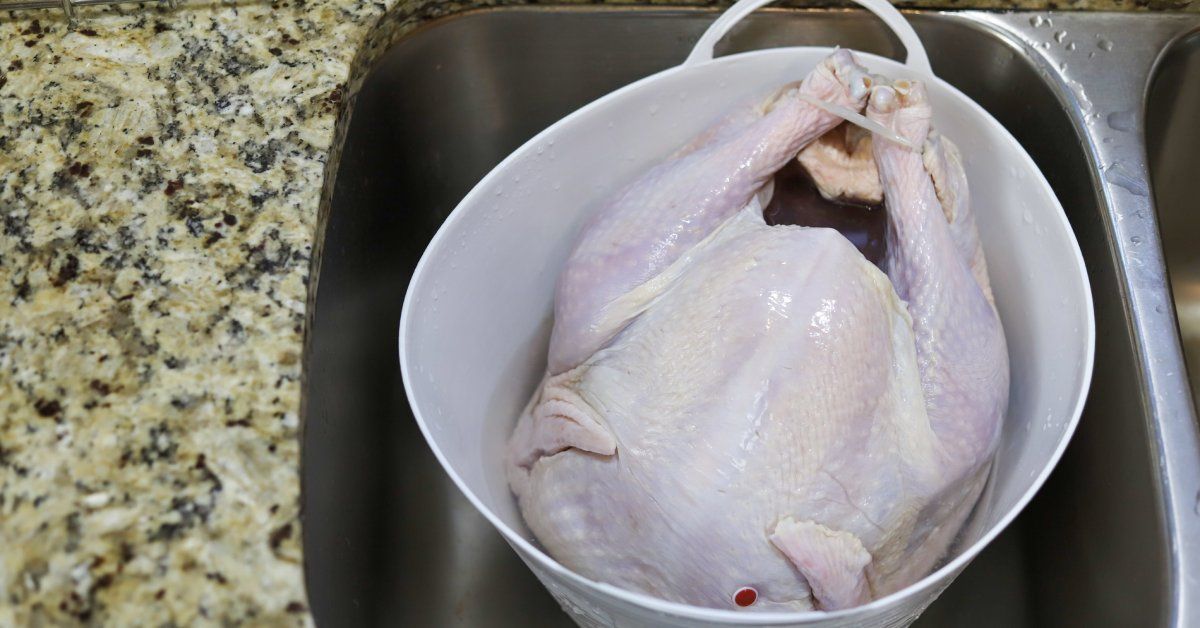 A raw turkey thawing in a white bucket in a stainless steel kitchen sink.