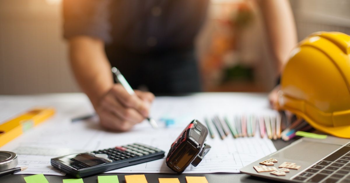 A contractor leans over a desk, marking up notes on a large sheet of paper, with tools and protective gear in the foreground.