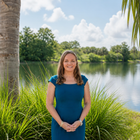 Woman in a blue dress standing by a lake with green plants and trees in the background