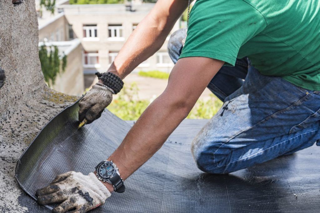 A man is kneeling down and using a shovel to spread a piece of asphalt on a roof.