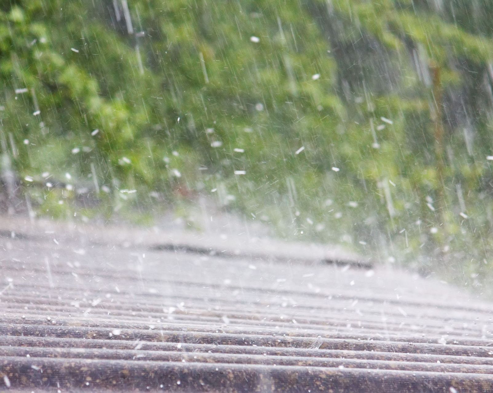 A close up of rain falling on a roof with trees in the background.