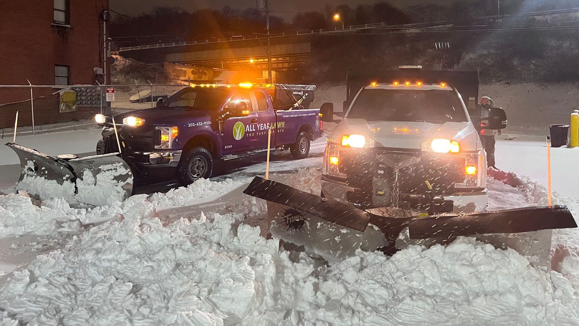 two snow plows are plowing snow in a parking lot at night .