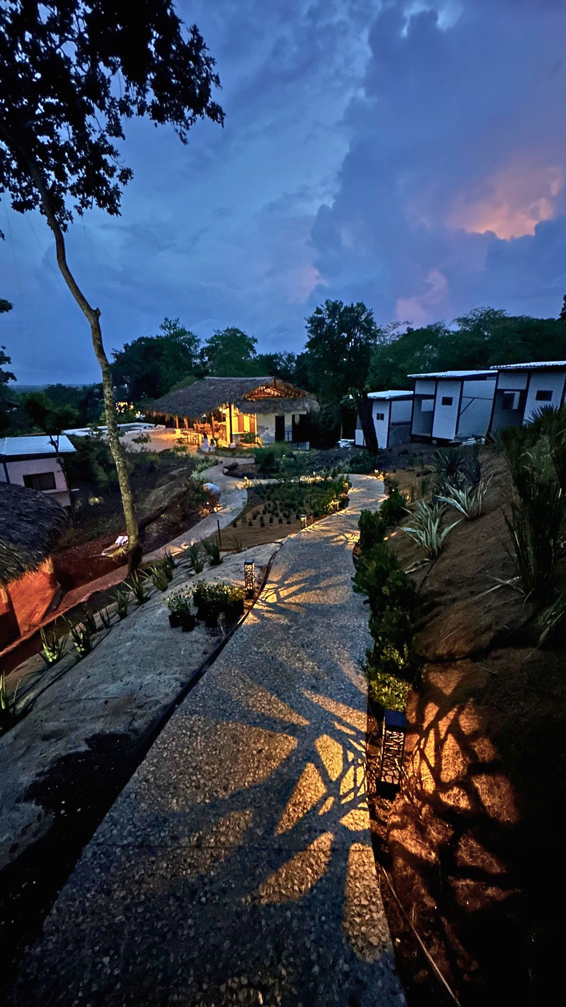 A twilight scene of a pathway leading to a lit-up building with a thatched roof. The sky is a mix of blues and pinks.