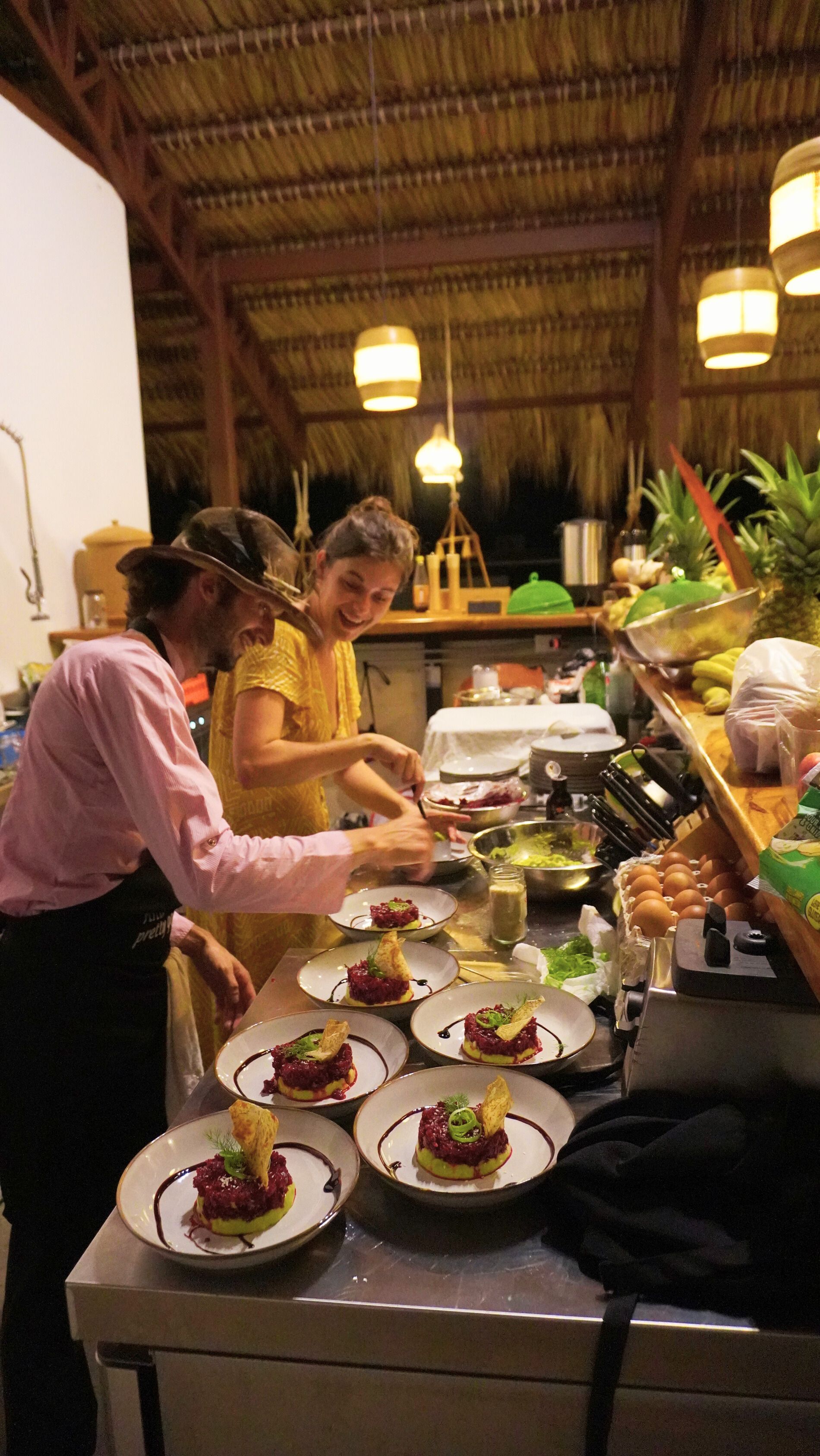 Two people plating food in a kitchen. The scene is dimly lit with hanging lights, featuring prepared plates.