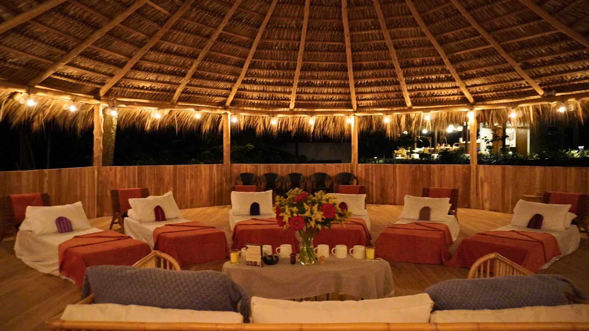 Circular, open-air hut at night, with several beds and pillows arranged around a central table with flowers.