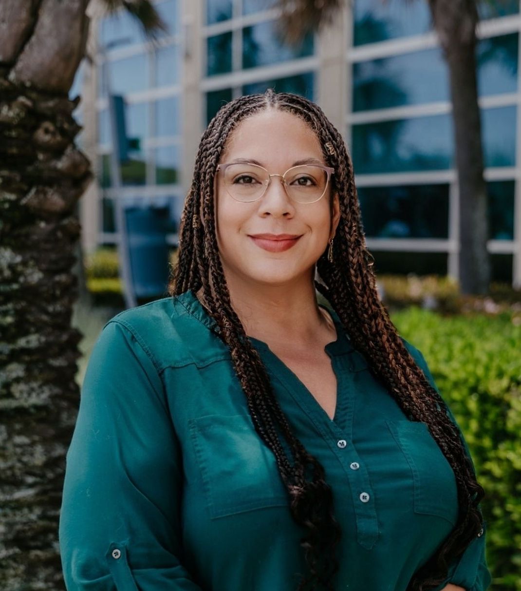 Woman with long, dark braids wearing glasses and a teal shirt, smiling slightly outside a modern building with palm trees.