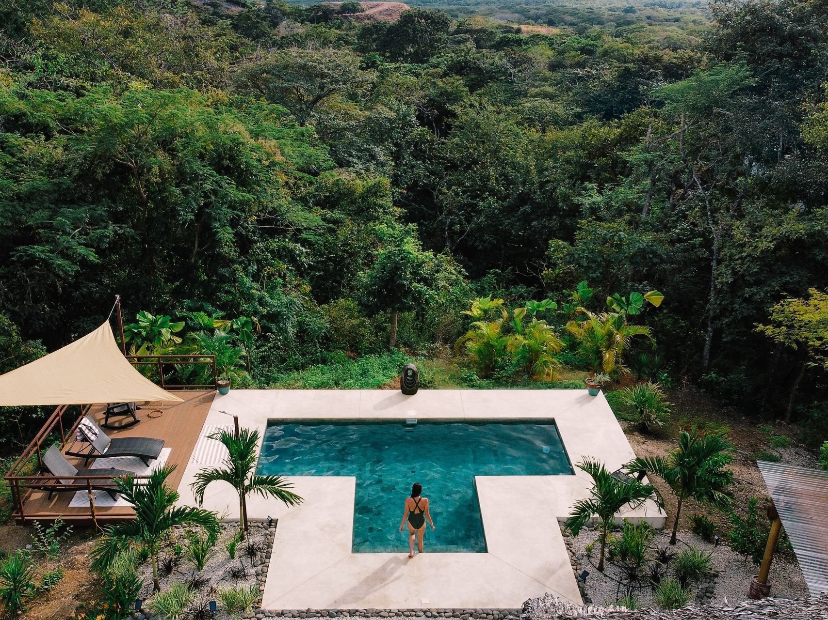 Woman standing at edge of a square pool, looking into the water. Lush green jungle surrounds the pool.