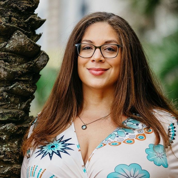Woman with long brown hair and glasses, smiling, leaning against a tree trunk. She wears a floral print top outdoors.