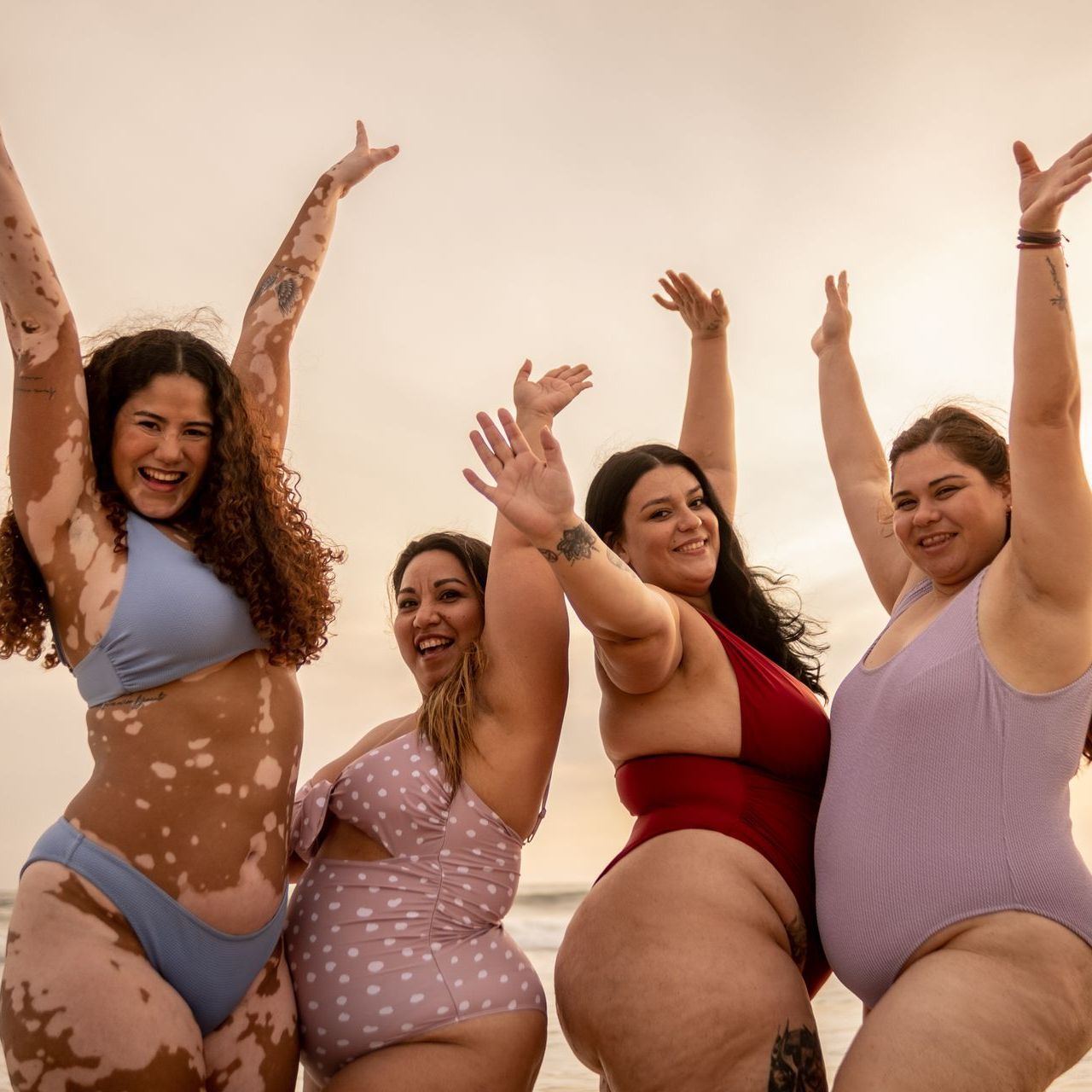 Four diverse women in swimwear raise their arms joyfully on a beach at sunset. One has visible vitiligo, others have tattoos.