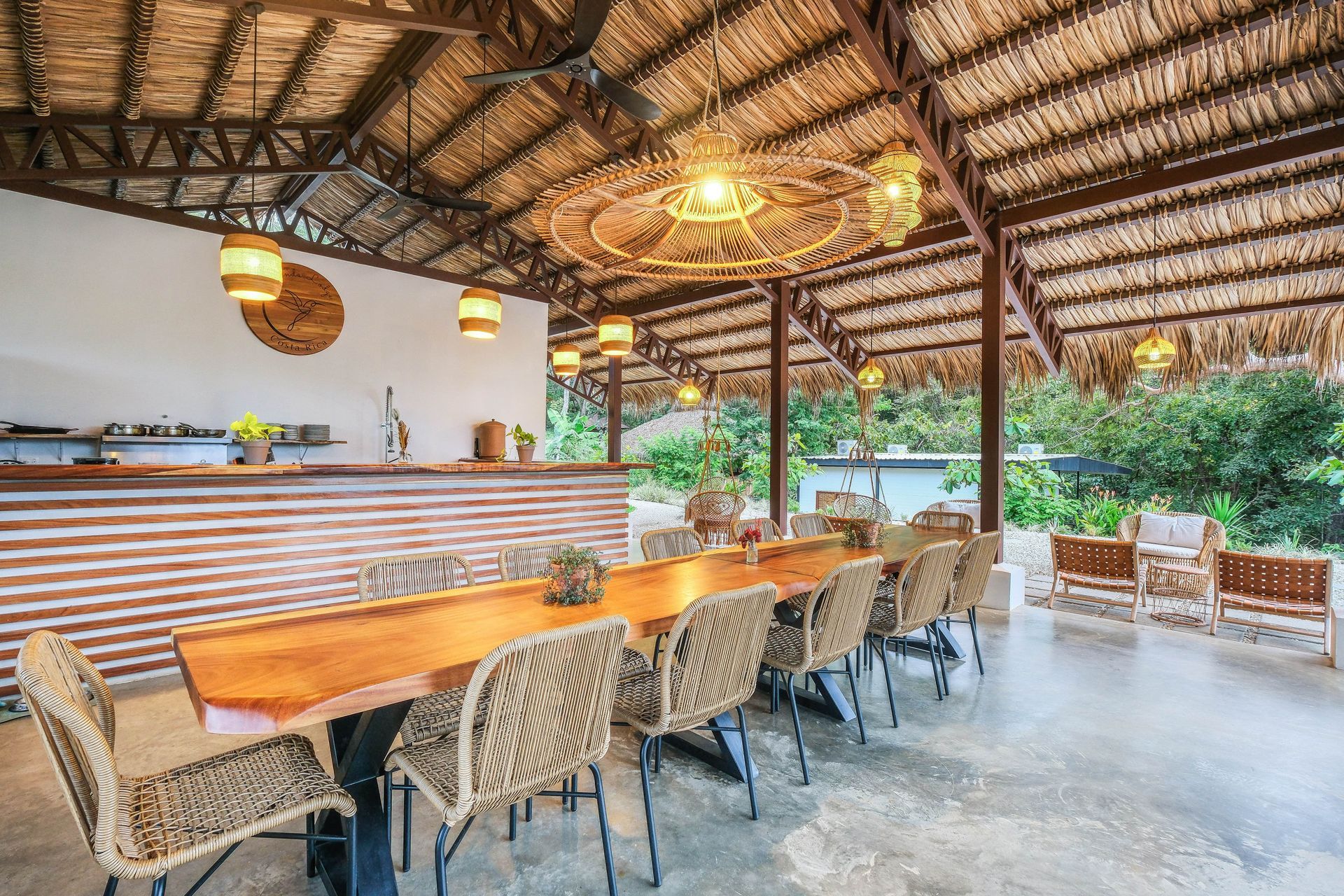 Outdoor dining area with a long wooden table, woven chairs, and a thatched roof.