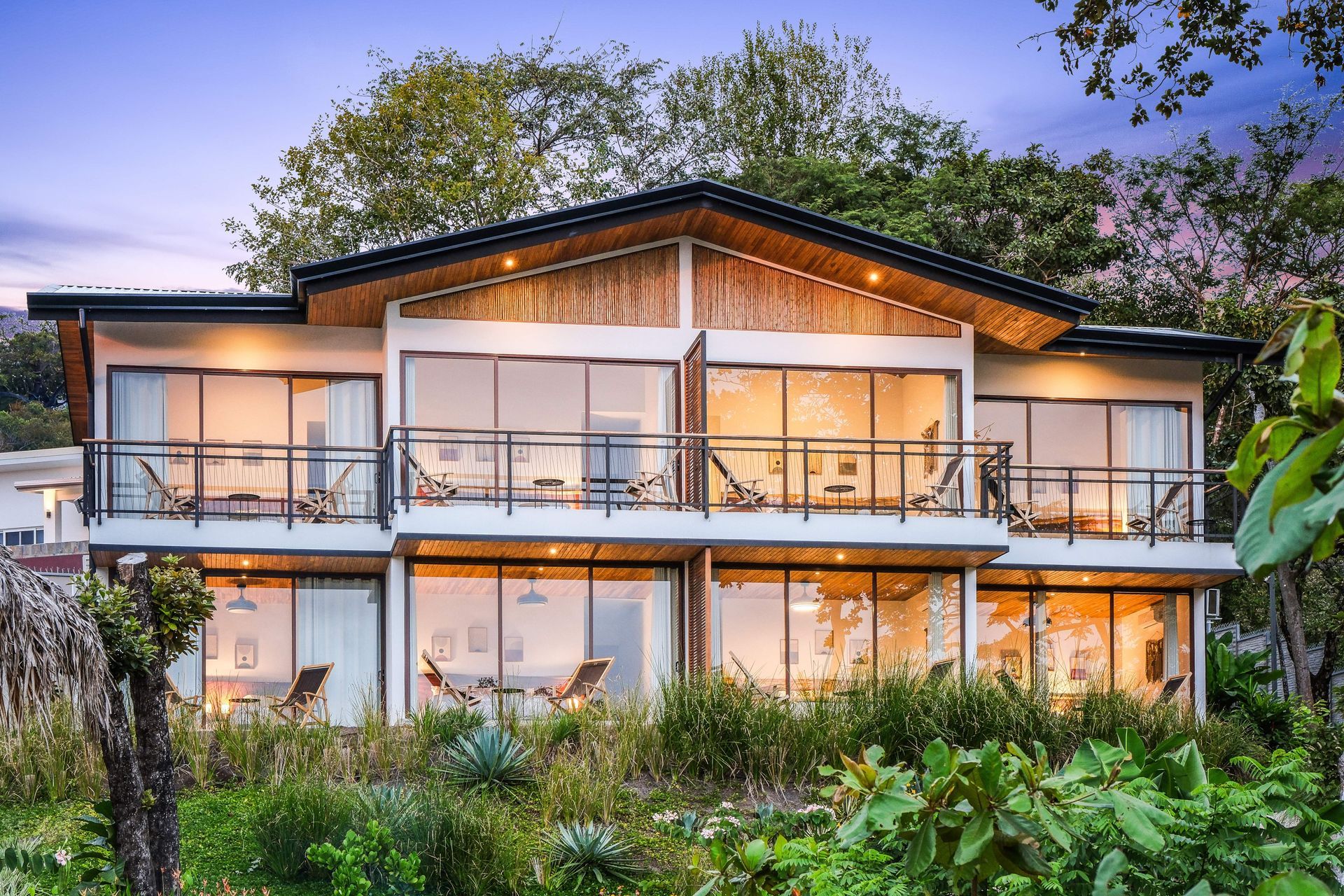 Two-story modern home with large glass windows and balconies, nestled in lush green foliage with a natural wood.