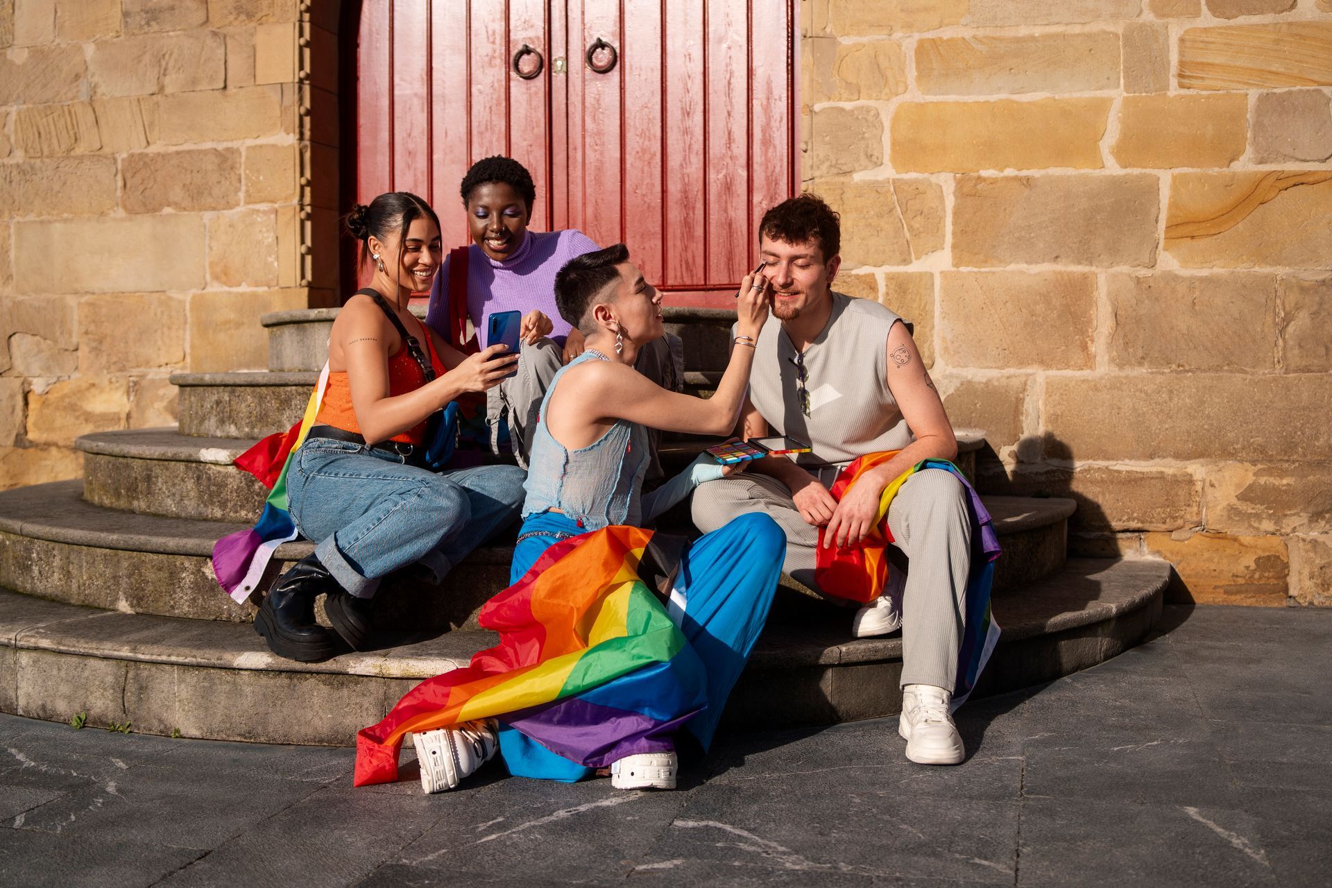 Four young people with rainbow flags sit on stone steps. They are taking photos and applying makeup.