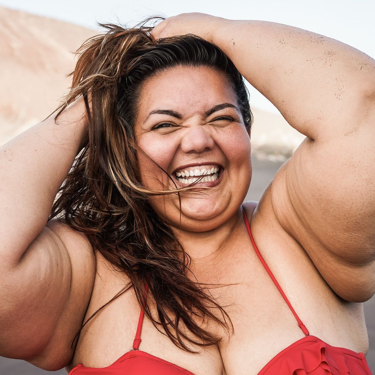 A smiling woman in a red swimsuit holds her hair up, laughing outdoors.