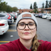 Une femme portant des lunettes et un bonnet prend un selfie sur un parking en souriant.