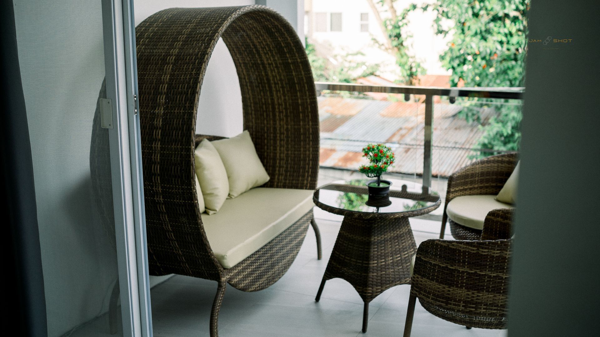 A balcony seating area featuring a round wicker sofa with beige cushions, a matching small table, and two wicker chairs.
