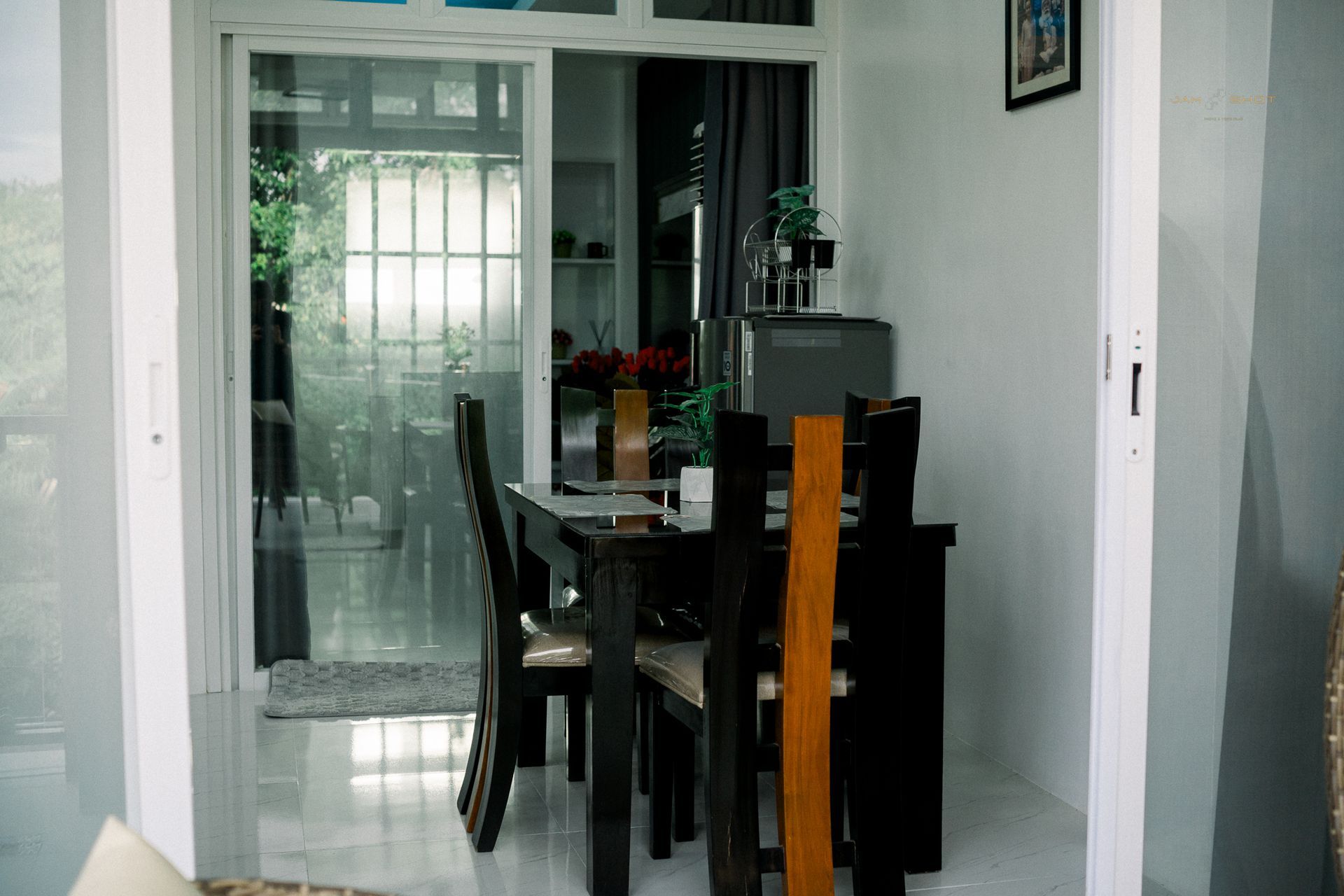 A dining table with chairs in a minimalist room, seen through an open doorway with a view to a glass-paneled outdoor area.