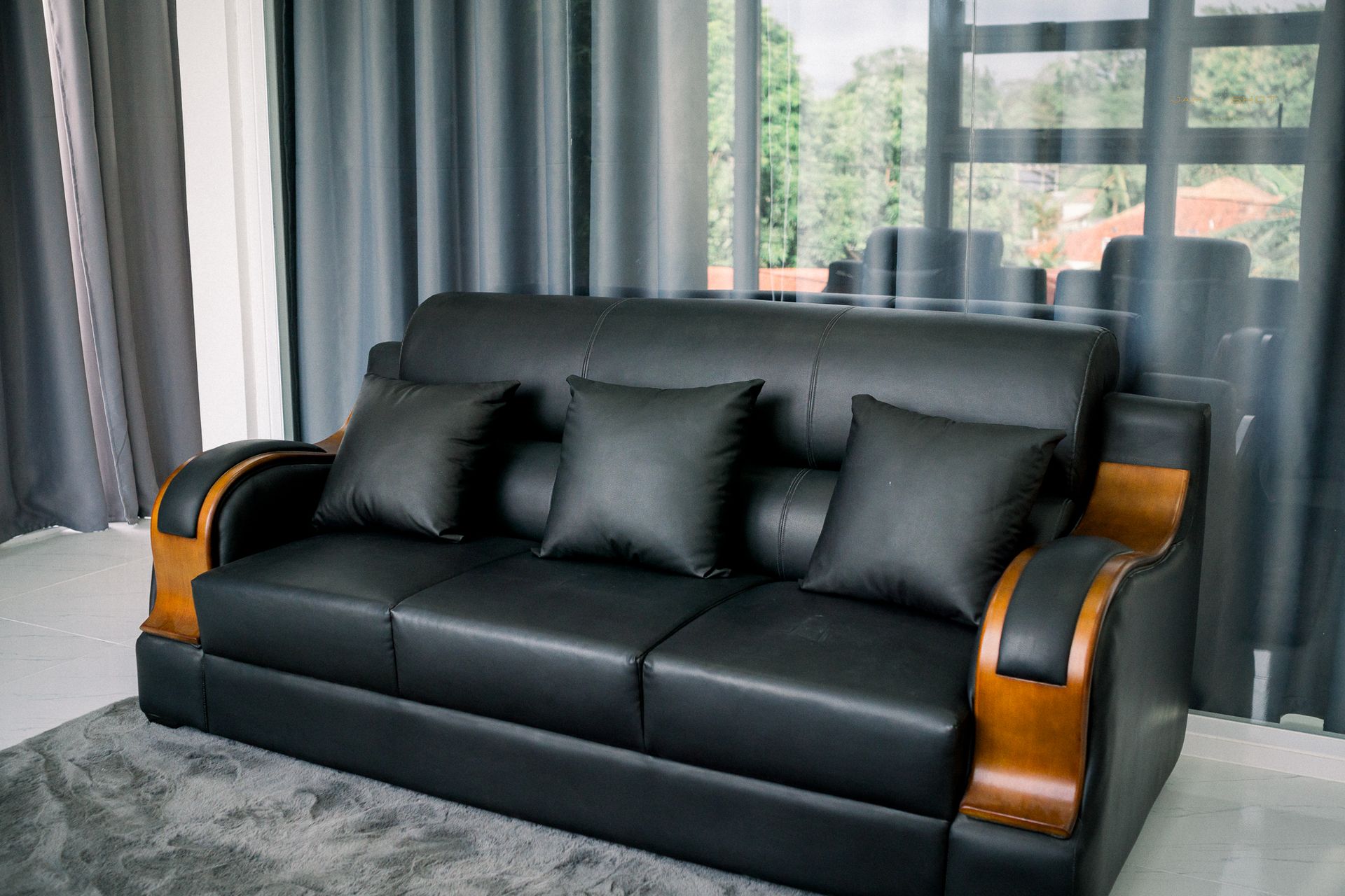 A black leather sofa with three matching cushions and wooden armrests, positioned in a room with grey curtains.