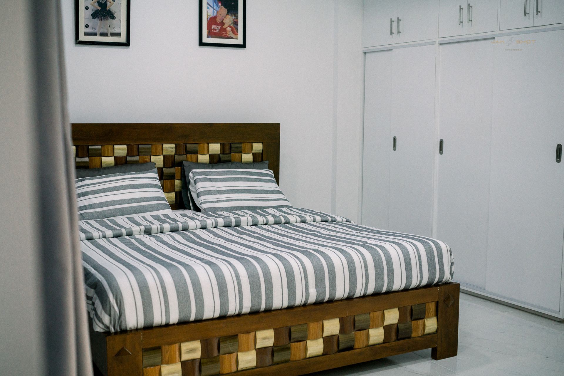 A modern bedroom with a wooden bed frame, grey and white striped bedding, white walls, and white built-in wardrobes.