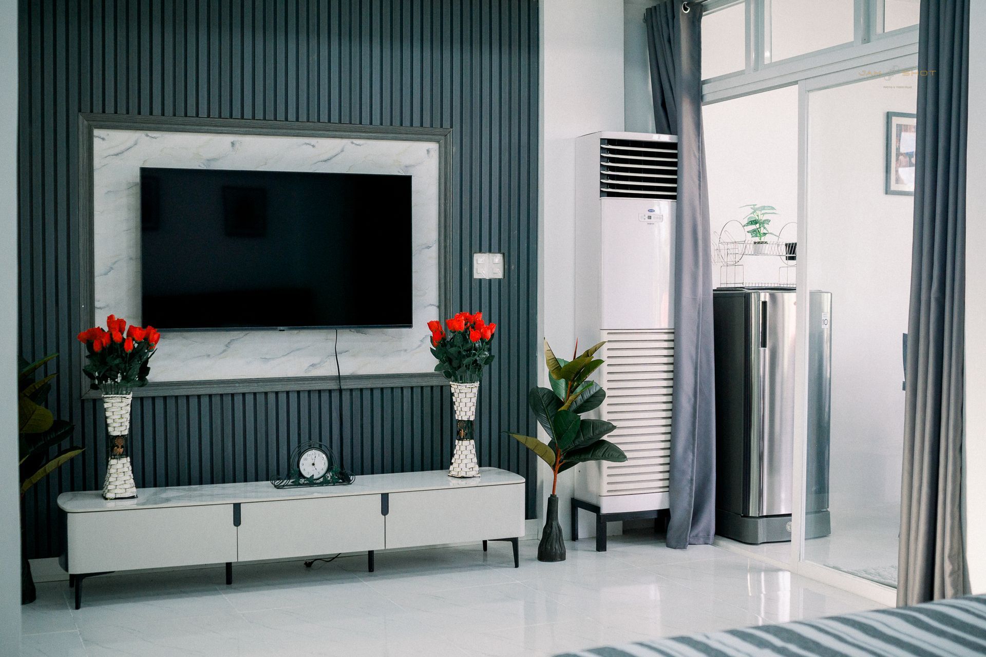 Modern living room with a wall-mounted TV on a slate-gray slatted wall, white media console, roses, and floor AC unit.