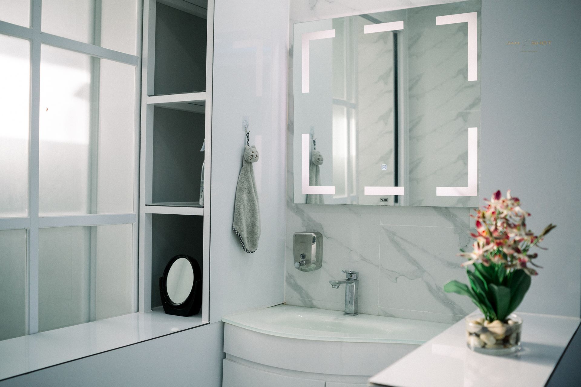 Modern white bathroom vanity with marble countertop, wall-mounted mirror, open shelving, and a small flower arrangement.