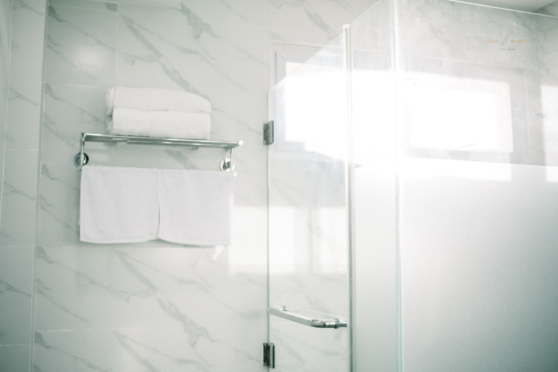 White towels hang on a chrome rack mounted on a marble-tiled wall next to a glass shower door.
