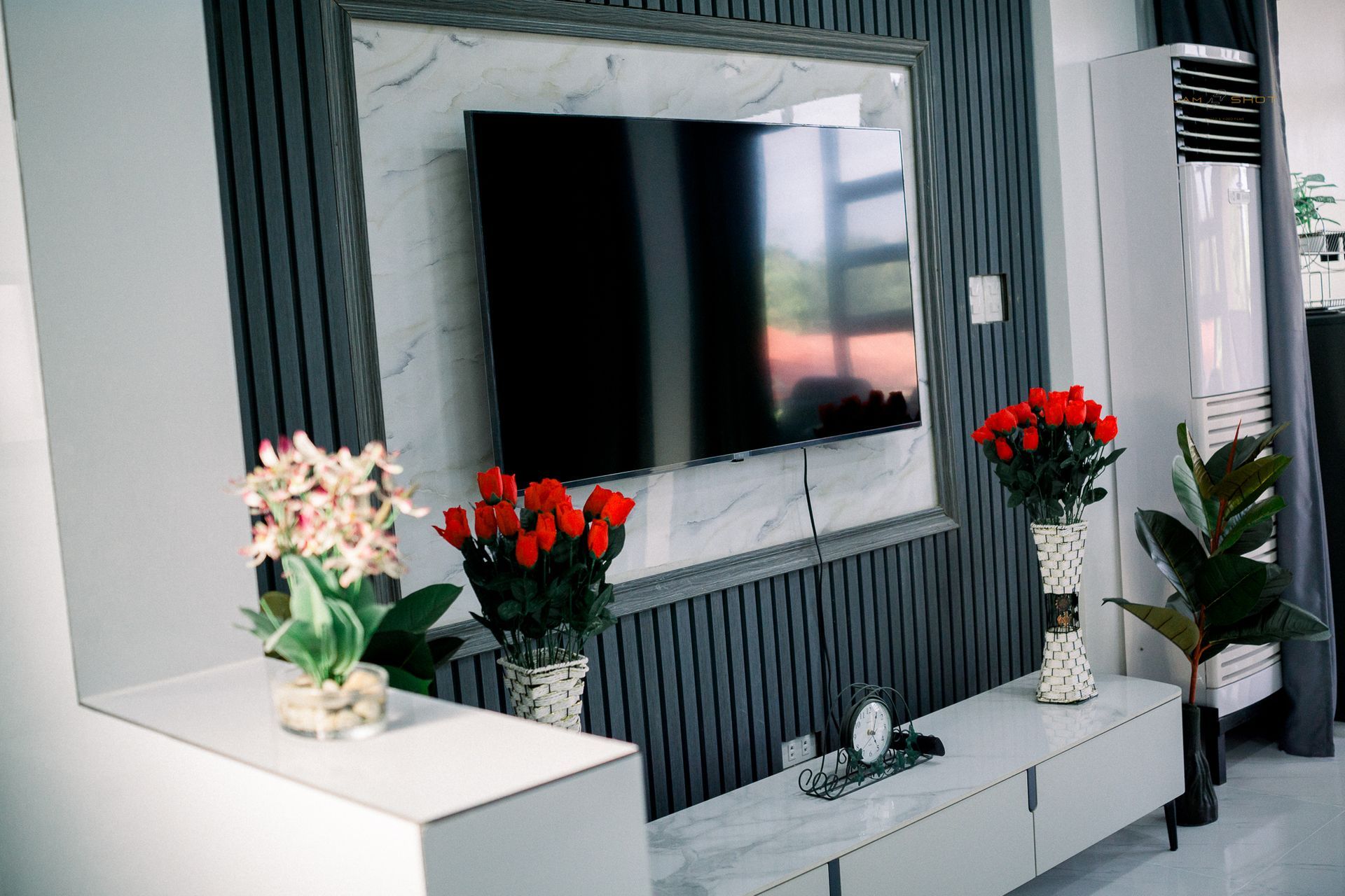 A wall-mounted TV framed by vertical slats and marble, with bouquets of red roses and an indoor plant on a white stand.
