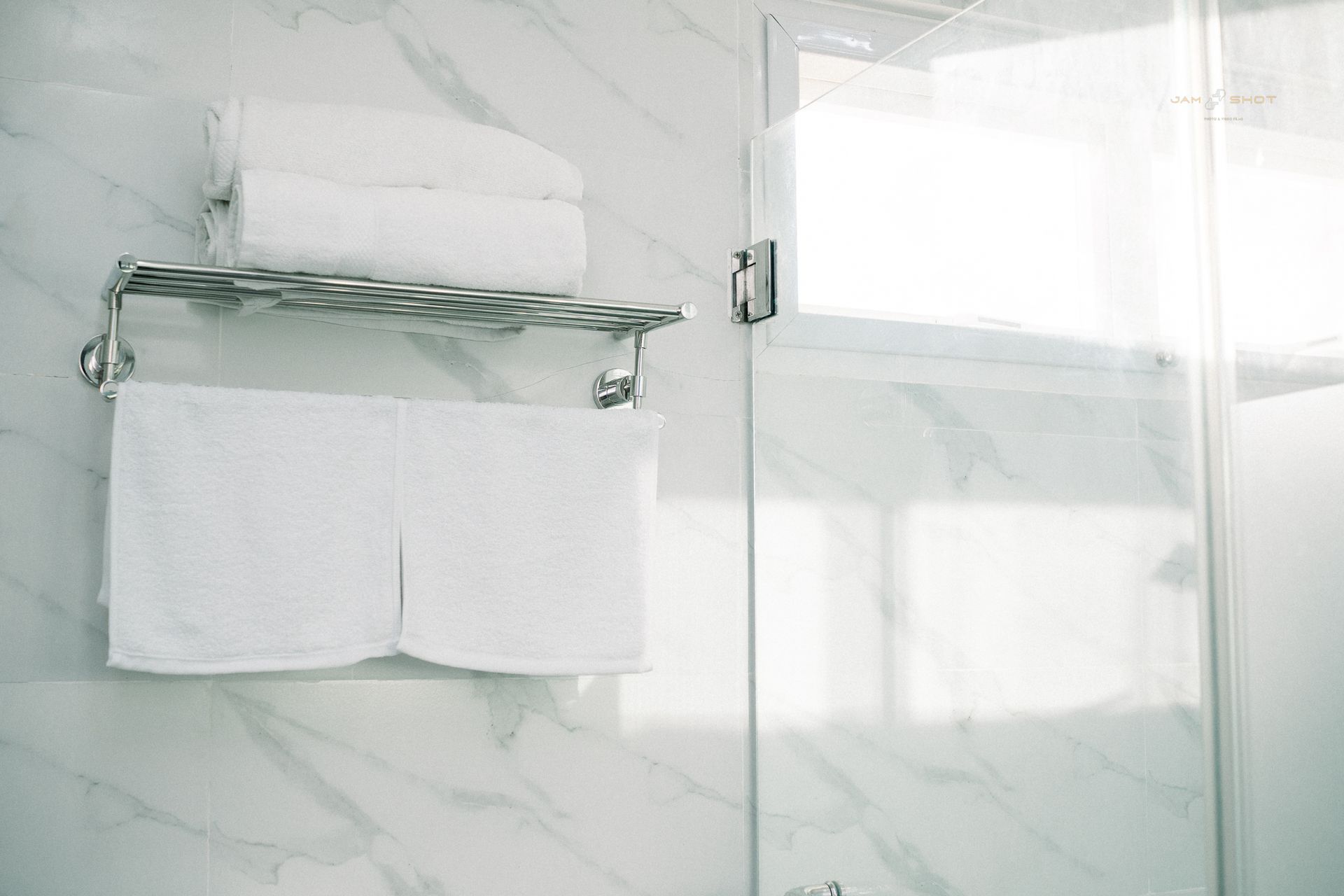 A silver towel rack holding folded white towels against a white marble-tiled wall next to a glass shower door.